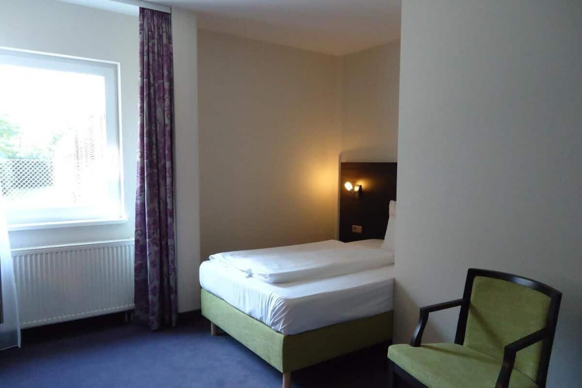 A small, neatly arranged hotel room with a single bed, a chair, a bedside table, and a window with curtains.