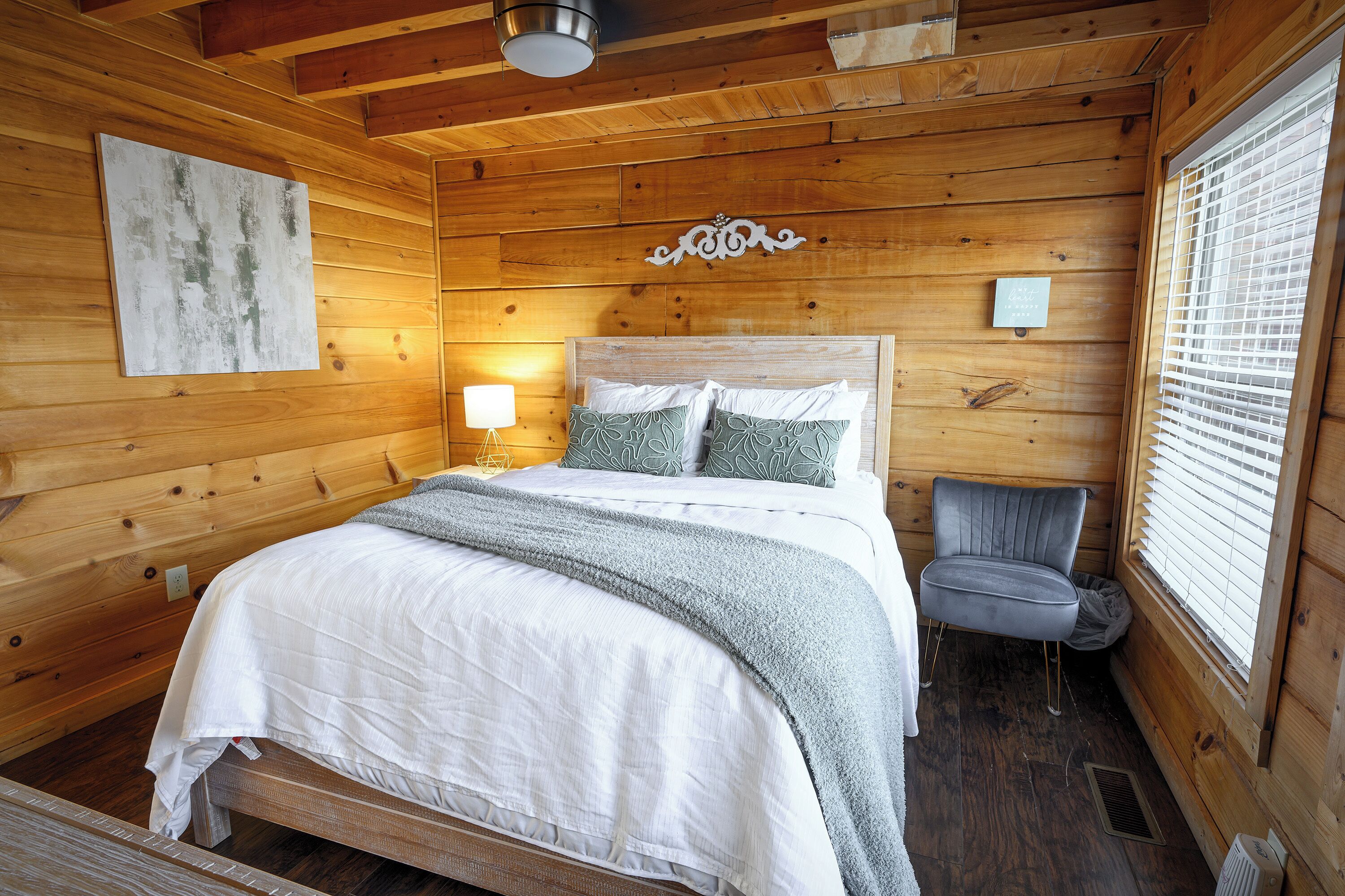 A wooden cabin bedroom with a bed, a chair, a lamp, and a window with blinds.