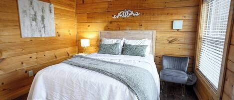 A wooden cabin bedroom with a bed, a chair, a lamp, and a window with blinds.