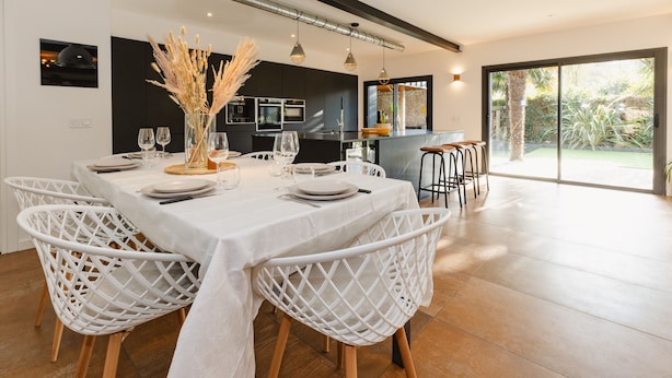 Un coin repas moderne avec une table blanche dressée pour six personnes, entourée de chaises en osier blanc. On aperçoit une cuisine à l’arrière-plan, avec des meubles de couleur sombre et une grande fenêtre.