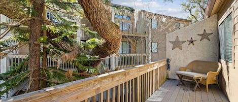 A wooden deck with a wicker sofa and a table, surrounded by trees and a building with star decorations.