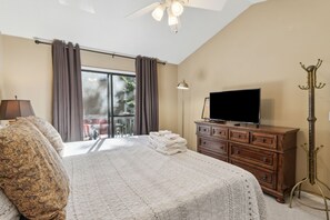 A hotel room with a large bed, a TV, a wooden dresser, a lamp, and a balcony with curtains.