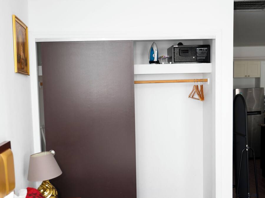 A hotel room with a bed, a nightstand with a lamp, a wardrobe, and a kitchenette in the background.
