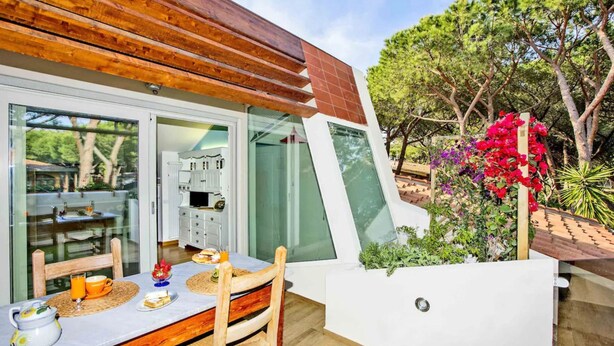 A modern outdoor dining area with a wooden table and chairs, a white cabinet, and a glass enclosure leading to a kitchen.