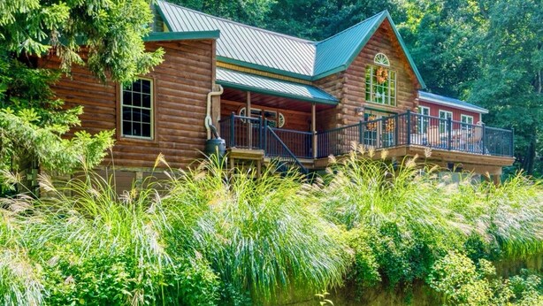 A log cabin with a porch and a deck, surrounded by lush greenery and a body of water.