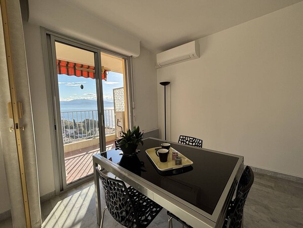 Un coin repas comprenant une table, des chaises, une plante en pot et une vue sur la mer, visible à travers une porte-fenêtre coulissante.