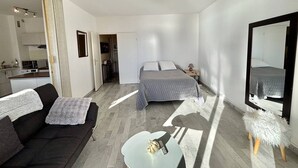 Une chambre d’hôtel moderne avec un lit, un canapé, une petite table, un miroir et, au fond, une kitchenette.