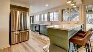 A modern kitchen with stainless steel appliances, a large island with stools, and a view of the outdoors through large windows. - Golf Course Home on 10th - Newly Renovated (Millersburg)