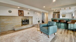 A modern living room with a fireplace, a blue armchair, and a kitchen area with bar stools. - Golf Course Home on 10th - Newly Renovated (Millersburg)