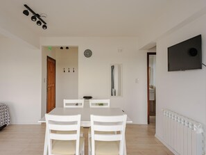 A dining area with a white table and chairs, a clock, a mirror, and a TV on the wall. - 1 bedroom apartment with sea view (Ushuaia - Tierra del Fuego)
