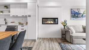 Una cocina moderna con muebles blancos, una zona de comedor con mesa de madera y sillas grises, una chimenea empotrada y un sofá gris en un salón con un cuadro en la pared.
