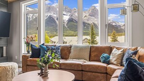 Ein Wohnzimmer mit braunem Ledersofa, einem runden Couchtisch und einem großen Fenster mit Blick auf die Berge.
