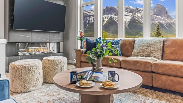 Ein modernes Wohnzimmer mit Kamin, großem Flachbildfernseher, braunem Ledersofa, rundem Couchtisch mit Blumenvase und Blick auf Berge durch große Fenster.
