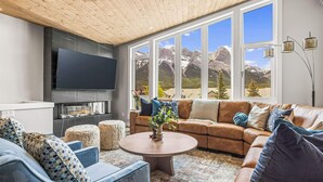 Ein modernes Wohnzimmer mit großem Fernseher, bequemen Sitzgelegenheiten, einem Kamin und Bergblick durch große Fenster.