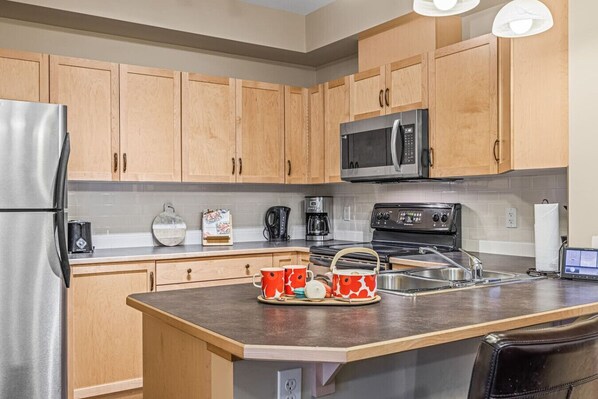 A modern kitchen with wooden cabinets, a stainless steel refrigerator, a microwave, a stove, and a sink.