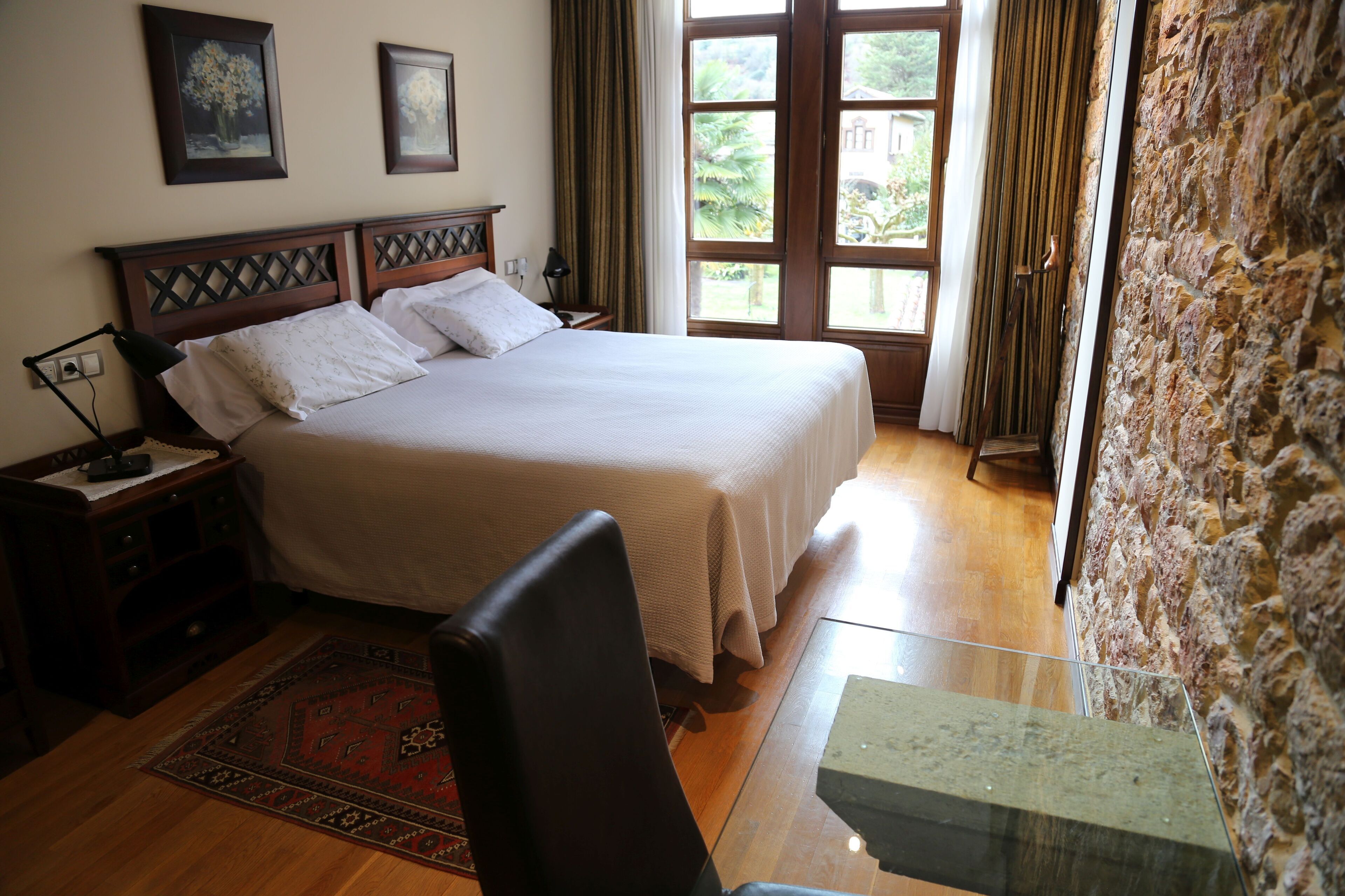 A bedroom with a large bed, wooden headboard, a nightstand, a desk, and a stone wall.