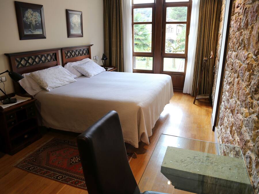 A bedroom with a large bed, wooden headboard, a nightstand, a desk, and a stone wall.