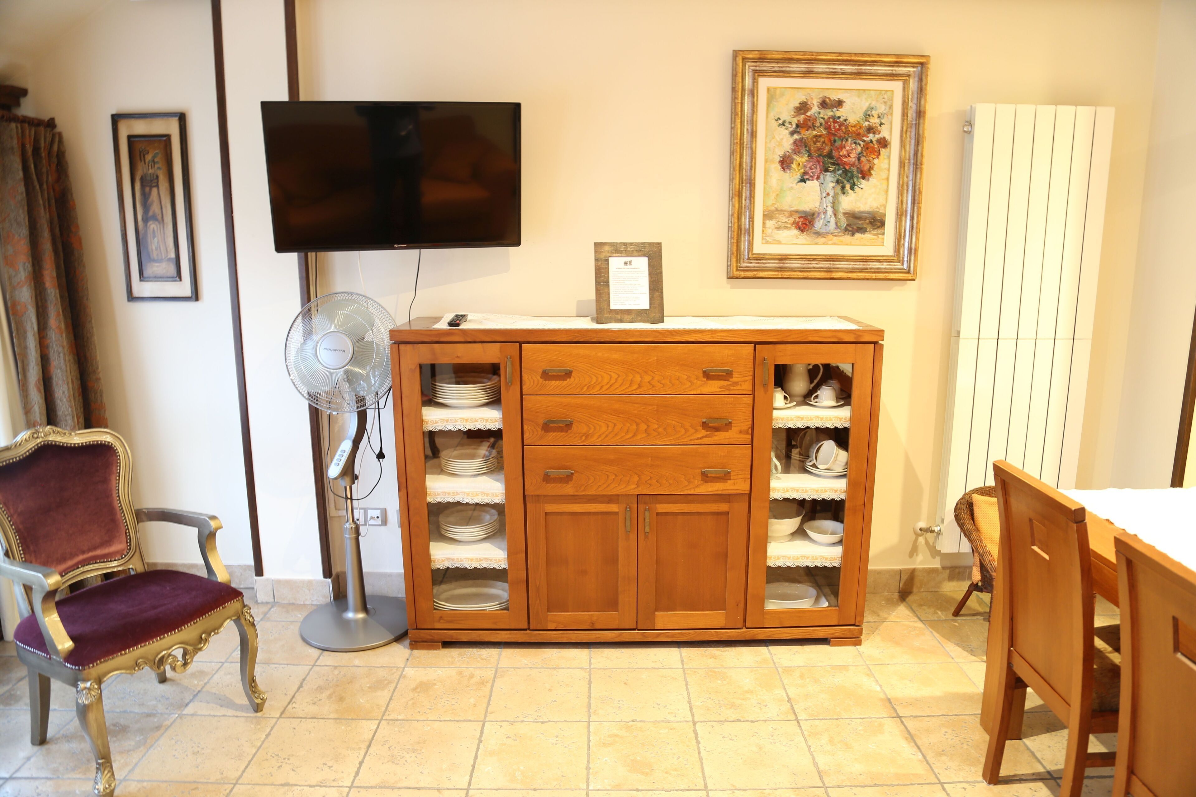 A room with a wooden cabinet with drawers, a television mounted on the wall, a framed painting, and a chair.