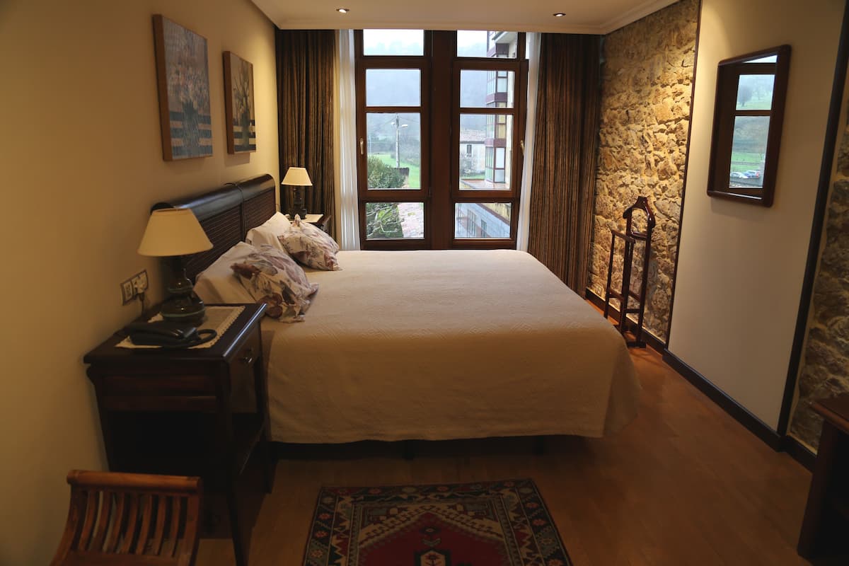 Habitación de hotel con una cama grande, una mesita de noche, una silla, una ventana con vistas a edificios y una pared de piedra.