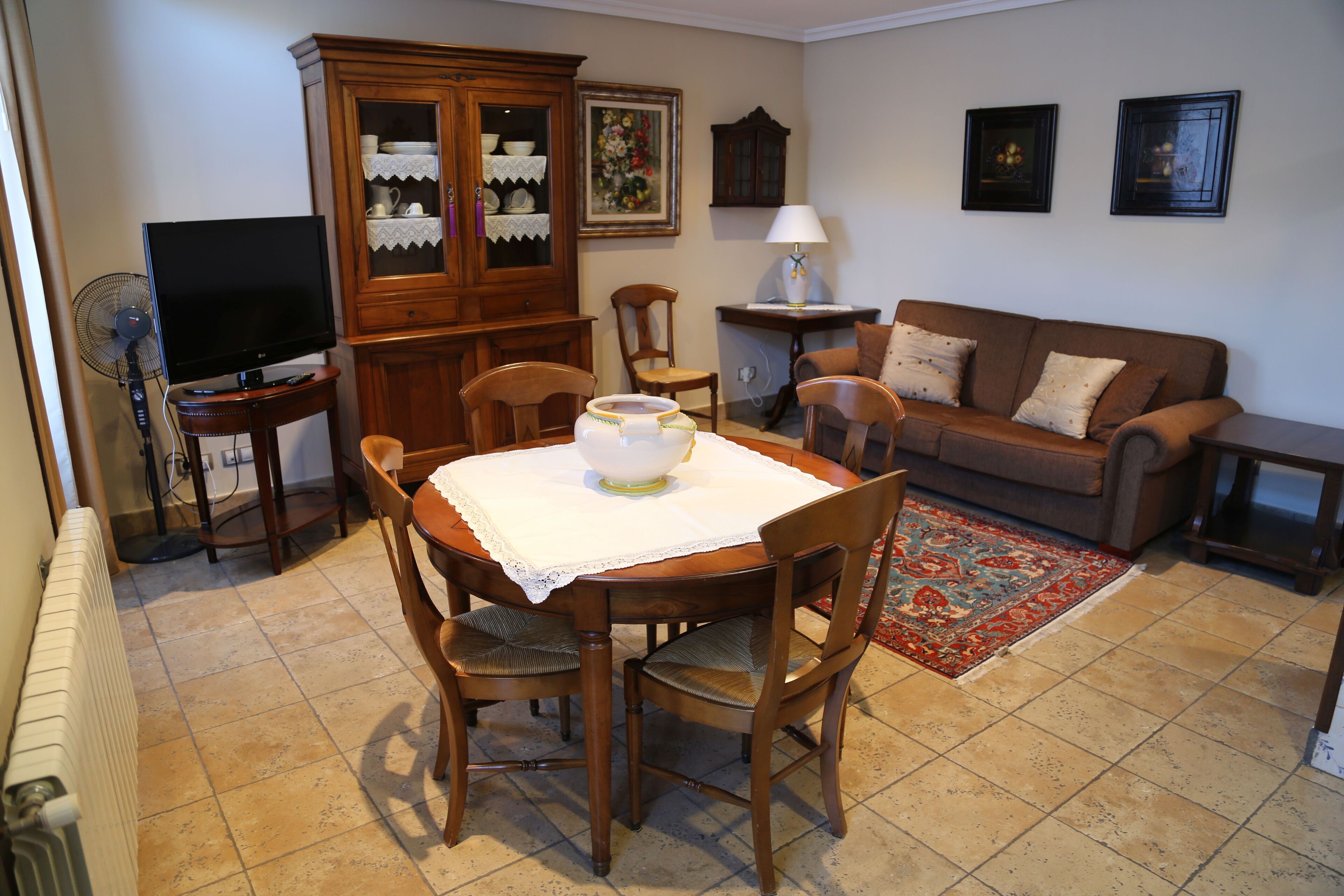 A dining area with a table set for four, surrounded by wooden chairs. A television is placed on a stand to the left.