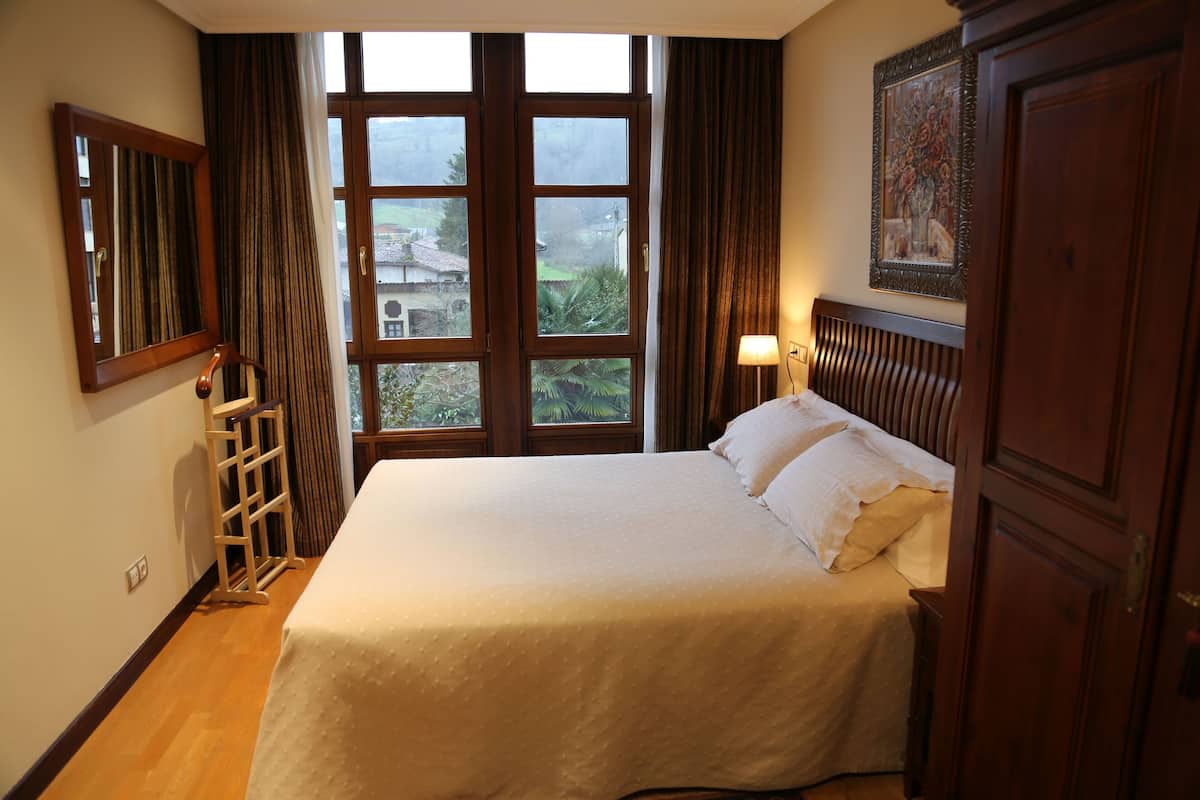 Una cama bien hecha con una colcha blanca, cabecera de madera y una mesita de noche con una lámpara. Se ve una ventana con cortinas, y un cuadro cuelga en la pared.