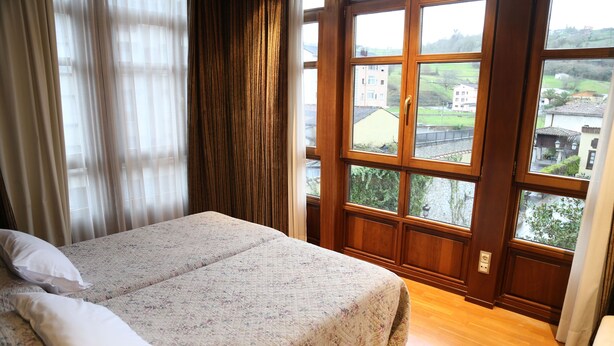 A bedroom with a bed, curtains, and a large window with a view of buildings and greenery. - La Pontiga (Pilona)