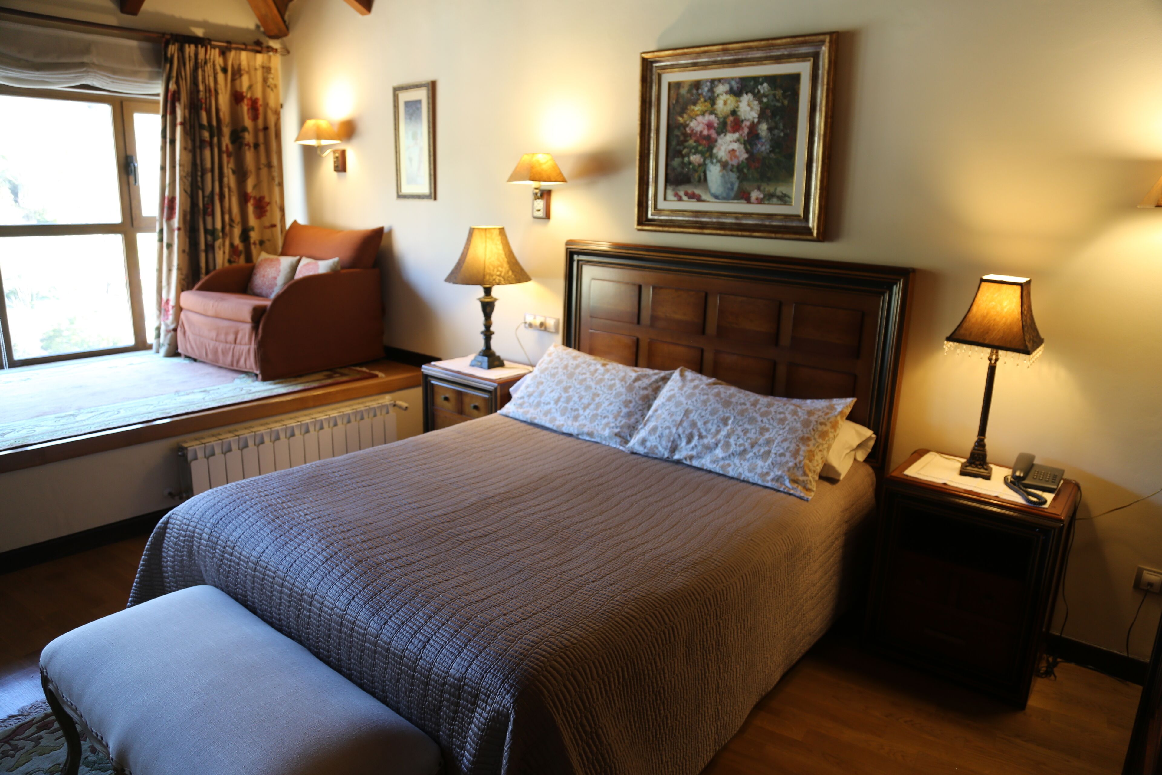 A neatly arranged hotel room with a large bed, a window seat with a chair, a bedside table with a lamp, and a painting on the wall.