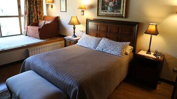 A neatly arranged hotel room with a large bed, a window seat with a chair, a bedside table with a lamp, and a painting on the wall.