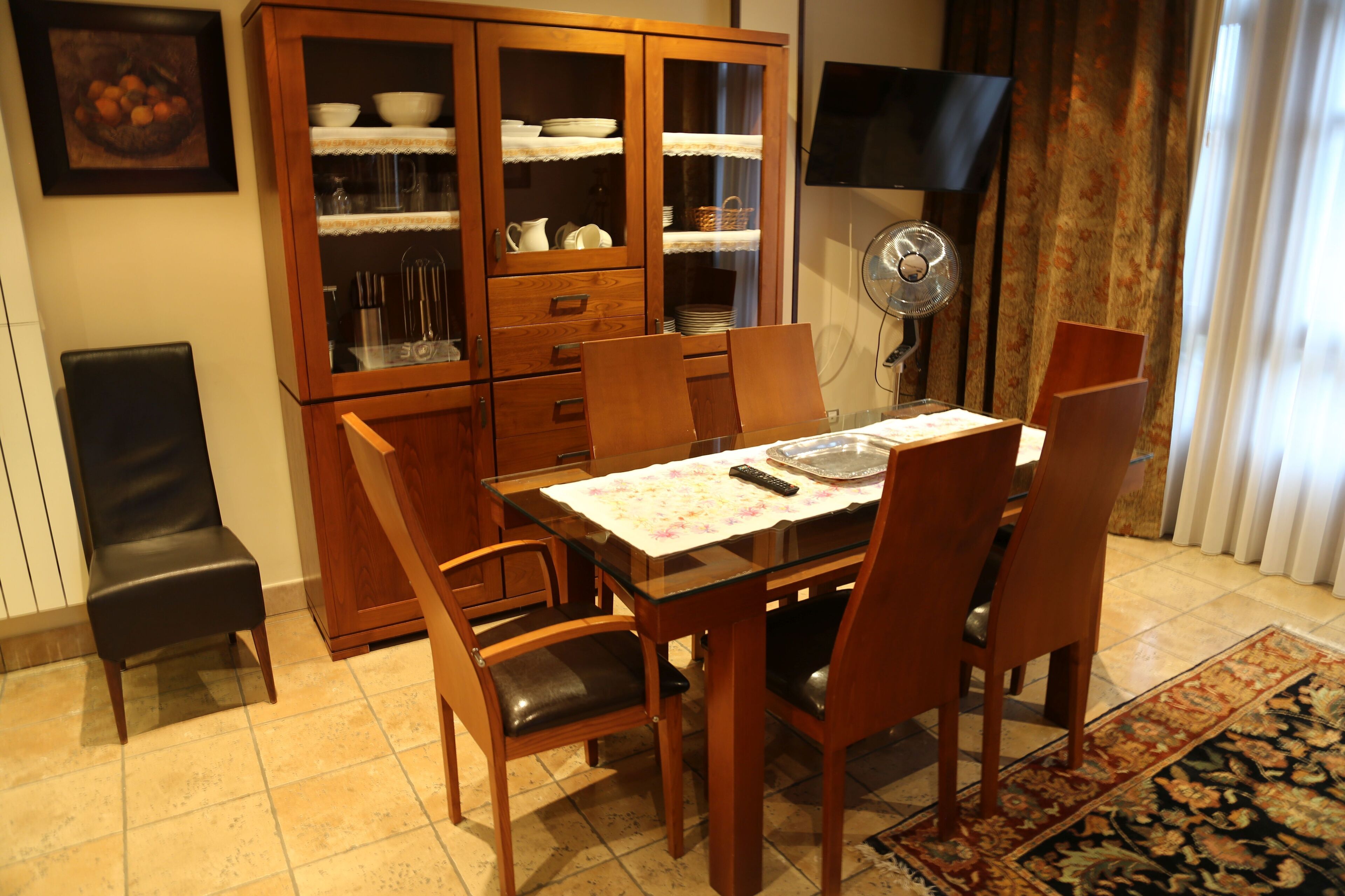 Un comedor con una mesa de vidrio, sillas de madera, un armario con vajilla y un televisor montado en la pared.
