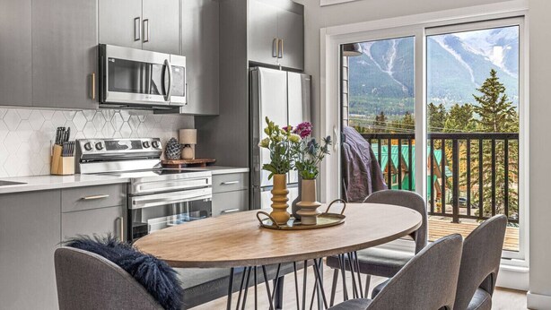 A modern kitchen with stainless steel appliances, a dining area with a wooden table and chairs, and a balcony with a mountain view. - Delightful 3BR Condo w/ AC+Stunning Mountain Views (Canmore)