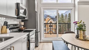 A modern kitchen with stainless steel appliances, a dining area with a table and chairs, and a large sliding glass door leading to a balcony with a view of a mountain and a house under construction. - Delightful 3BR Condo w/ AC+Stunning Mountain Views (Canmore)