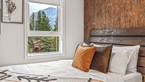 A bedroom with a bed, a window with a mountain view, and a wooden headboard. - Delightful 3BR Condo w/ AC+Stunning Mountain Views (Canmore)