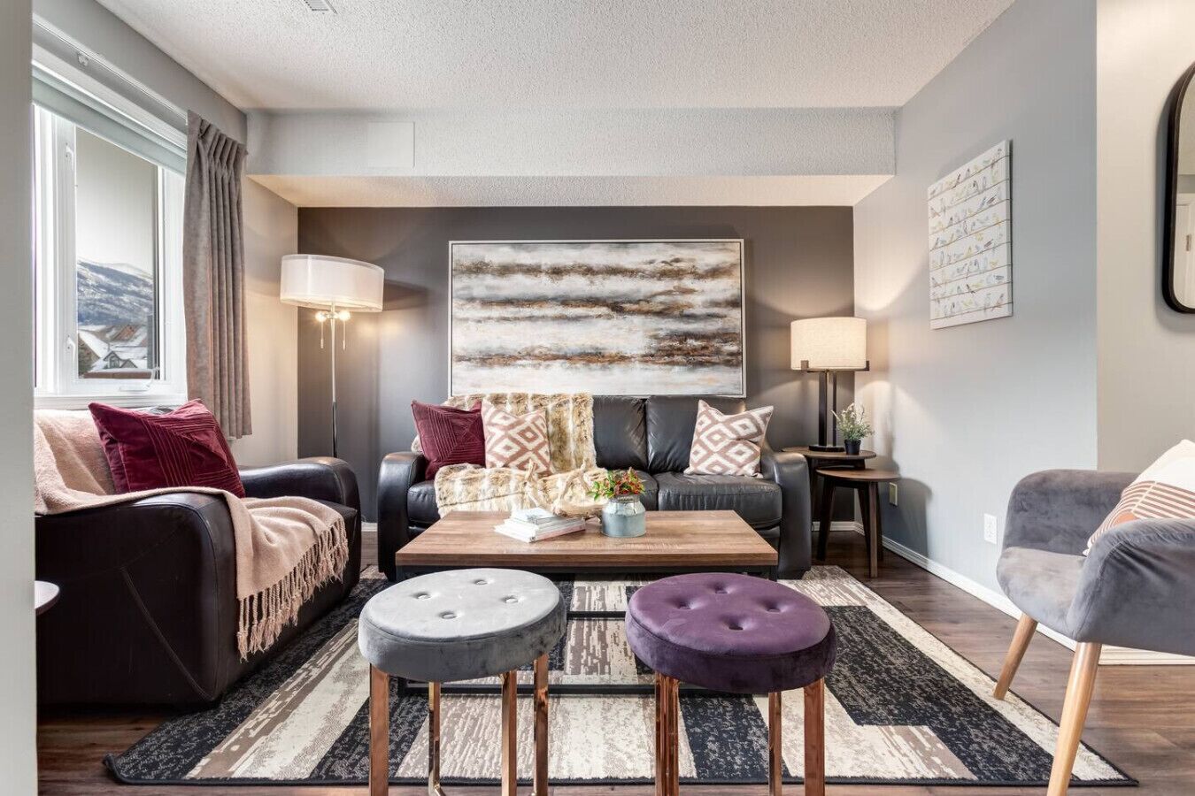 Un salon moderne avec un canapé, une table basse et deux poufs. Un grand tableau abstrait orne le mur, et une fenêtre est agrémentée de rideaux.
