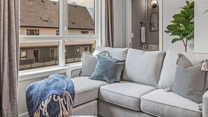 Een moderne woonkamer met een grijze modulaire bank, een blauwe plaid en een groot raam met uitzicht op een gebouw.