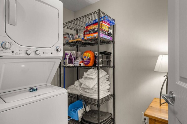 A laundry room with a washing machine, shelves with cleaning supplies, and a small table with a lamp.