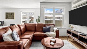 Una sala de estar con un sofá de cuero marrón, una mesa de centro redonda, un televisor y una ventana con vistas a edificios y montañas.