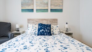 Un dormitorio con una cama, dos mesitas de noche, una lámpara, una silla y tres cuadros con escenas marinas.