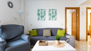 Una sala de estar con un sofá gris, dos cojines, una mesa de centro blanca con una bandeja tejida y una planta en maceta.