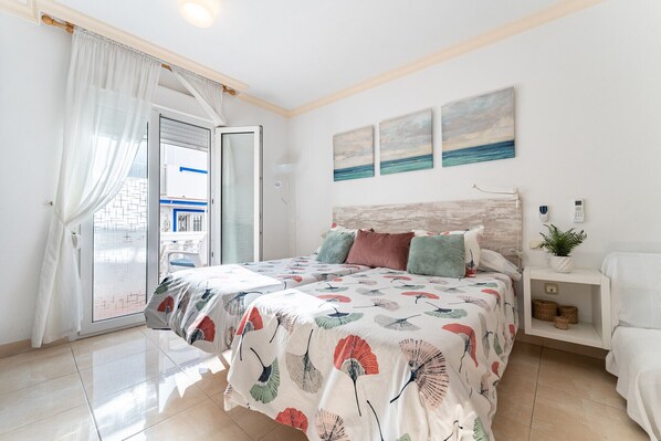 A bedroom with a bed, a bedside table, a nightstand, a potted plant, and a view of the outside.