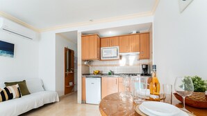 Una cocina moderna con armarios de madera, microondas y cafetera. Hay un comedor con una mesa preparada para dos personas, un sofá y una planta.