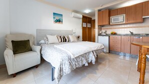 Un dormitorio con cama, una silla, una kitchenette y un ventilador de techo.