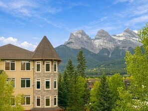 A multi-story building with a stone facade and a pitched roof, surrounded by lush greenery and mountains in the background.