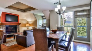 A modern living room with a fireplace, a dining area, and a large window offering a mountain view. - Superb 2BR Condo w/ Charming Mountain View & Pool (Canmore)