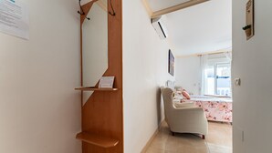 A hallway with a wooden door, a small shelf with a sign, and a chair in a room with a bed and a window.