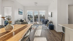 Uma sala ampla com uma mesa de jantar de madeira, um sofá e uma varanda com vista para árvores e montanhas.