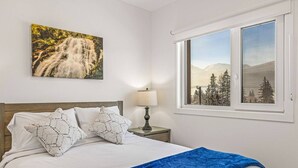 Quarto com cama, almofadas, mesa de cabeceira com luminária, janela com vista para montanhas e um quadro de uma cachoeira.