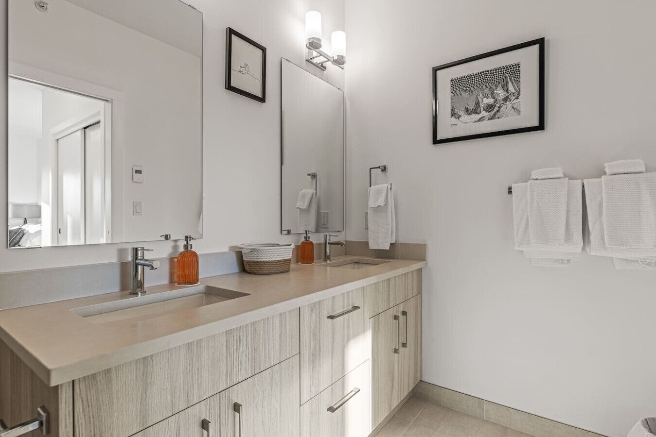 Une salle de bain moderne avec un double vasque, des meubles en bois, un grand miroir et des œuvres d’art encadrées accrochées au mur.