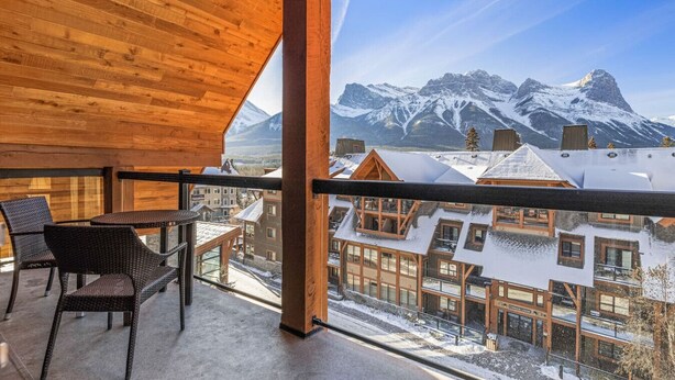 Un balcón de madera con una mesa y sillas, con vistas a un paisaje montañoso nevado y edificios.