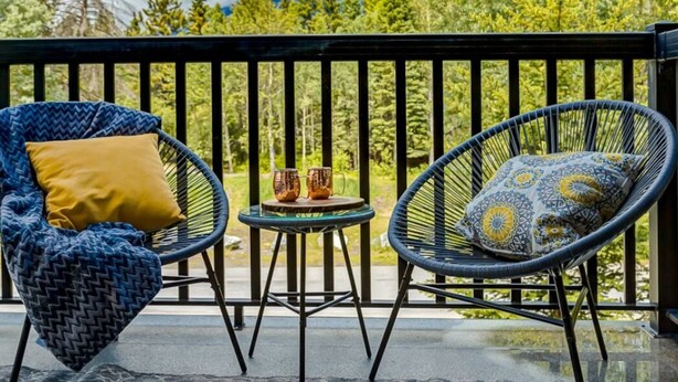A balcony with two wicker chairs and a small table, overlooking a forested area with mountains in the background.