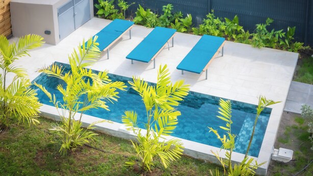 Una piscina con sillas reclinables azules, rodeada de follaje y un muro de ladrillo.